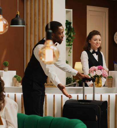 Helpful concierge assists elderly guest for check in, giving room key card while bellhop carries his luggage. Retired person waits at front desk while receptionist verifies identity with passport.